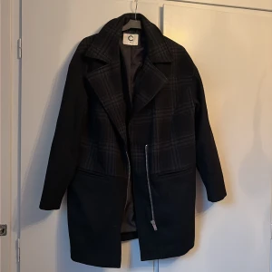 Svart rutig kappa från C, strl 36 - Snygg svart kappa från C i storlek 36 med rutigt mönster upptill och stilren design. Jackan har bred krage, långa ärmar och en cool sned dragkedja framtill. Materialet känns som ull och polyester, perfekt för höst och vinter.