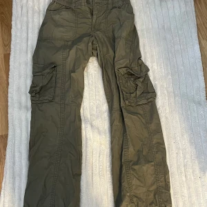 Khakigröna cargobyxor från Gina Tricot - Säljer ett par khakigröna cargobyxor från Gina Tricot med stora fickor på sidorna och bak. Byxorna har normal passform, elastisk midja och är tillverkade i ett lätt bomullsmaterial. Perfekta för en avslappnad och trendig look.