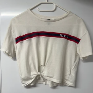 Vit croppad topp med NYC-tryck från H&M - Säljer en vit croppad topp från H&M i storlek XS. Toppen har korta ärmar, ett rött och marinblått randigt band med NYC-tryck över bröstet och en snygg knytning framtill. Tillverkad i mjuk bomull, perfekt för en chill och sportig look.