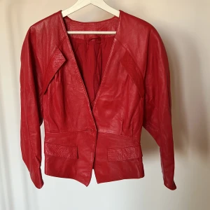 Röd skinnjacka med bred krage - Cool röd skinnjacka med breda axlar och markerad krage. Jackan har en knappstängning framtill och fickor med snyggt ormskinnsmönster på locken. Modellen är figursydd och har långa ärmar. Perfekt statement-plagg för dig som vill sticka ut.