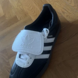 Adidas Samba LT svartvita sneakers - Klassiska Adidas Samba sneakers i svart skinn med vita detaljer och de ikoniska tre ränderna på sidan. Skon har vit plös med Adidas-logga, snörning och gummisula med mönster undertill. Perfekt för dig som gillar retrostil och sportig vibe.