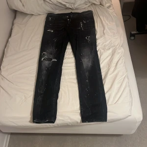 Svarta Dsquared2 Cool Guy jeans - Säljer ett par svarta Dsquared2 Cool Guy jeans med slitna detaljer, hål och färgstänk för en edgy look. Jeansen har klassisk femficksdesign, raka ben och en snygg patch på bakfickan. Tillverkade i Italien av bomull. Den är äkta.