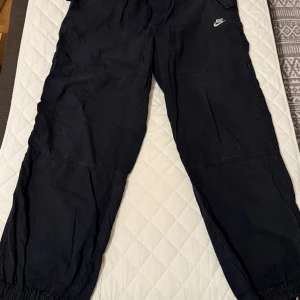 Svarta cargopants  - Svarta cargopants från Nike i storlek XL med broderad logga på vänster ficka. Byxorna har elastiska muddar vid bensluten, flera fickor och är tillverkade i bomull. Perfekt för en chill streetwear-look.