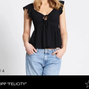 Topp Felicity Lager157 - Supersöt svart blus från Lager 157 med volangärmar och knytning framtill. Blusen har v-ringad hals och en lätt rynkad, flowy passform. Materialet är mjukt och känns som bomull, perfekt för att styla med jeans eller kjol. Storlek XS.
