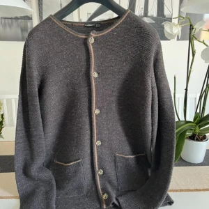 Mörkgrå kofta från Livergy, M - Snygg mörkgrå stickad kofta från Livergy med beige kant och dekorativa knappar framtill. Koftan har två praktiska fickor och långärmade ärmar. Perfekt för dig som gillar klassisk stil med en twist.