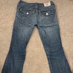 True Religion blå jeans med broderade fickor - Snygga blå jeans från True Religion med ikoniska broderade bakfickor och silverfärgade knappar. Modellen har låg midja och är bootcut, klassisk tvättad denim och tydliga kontrastsömmar. Perfekt för dig som gillar street och premium denim. Omsydda från en storlek 29 till en storlek 25/26 ( inte hål mellan skrevet utan sytt)