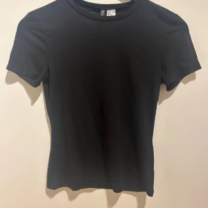 Svart basic t-shirt  - En svart, klassisk t-shirt från Divided i storlek S. Modellen är enkel med rund halsringning och korta ärmar. Tillverkad i mjuk bomull som känns skön mot huden. Perfekt att matcha med jeans eller kjol för en clean look.