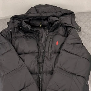 Svart pufferjacka från Polo Ralph Lauren - Svart pufferjacka från Polo Ralph Lauren med röd broderad logga på bröstet. Jackan har huva, tvåvägsdragkedja och en bröstficka med dragkedja. Materialet är syntet och jackan är quiltad för extra värme. Perfekt för kalla dagar.