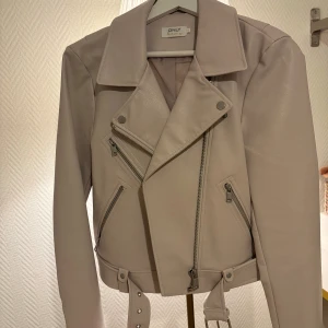 Beige skinnjacka från ONLY - Säljer en beige skinnjacka från ONLY med coola detaljer som flera dragkedjor och bälte i midjan. Jackan har klassisk krage och långärmade ärmar, perfekt för dig som gillar en edgy stil. Passformen är normal och materialet känns mjukt och snyggt.
