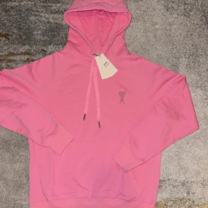 Rosa hoodie från Ami Paris - Säljer en snygg rosa hoodie från Ami Paris med broderad logga på bröstet. Tröjan har huva med snörning, känguruficka och ribbade muddar. Perfekt för dig som gillar streetstyle och vill sticka ut med färg.