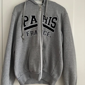 Grå hoodie med Paris-tryck och dragkedja - Zip up köpt några år sen i Paris i en turist affär. Är i storlek M. Är i ganska tjockt material och är marin blå inuti luvan. Skriv om du har frågor💗