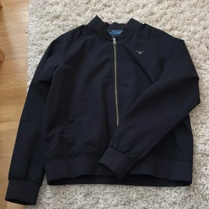 Svart bomberjacka från GANT - Svart bomberjacka från GANT.. Jackan har dragkedja framtill, två sidofickor och broderad GANT-logga på bröstet. Insidan är ljusblå med stor GANT-tryck. Skriv vid minsta fråga! Väldigt bra skick då den knappt har blivit använd