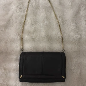 Svart axelväska med guldkedja - Svart axelväska i skinnmaterial med guldfärgade detaljer och kedja. Väskan har en rektangulär form, magnetlås och minimalistisk design. Perfekt storlek för det viktigaste och många rymliga fack i en liten väska. Hyfsat mycket slitage därav priset som också kan diskuteras💗
