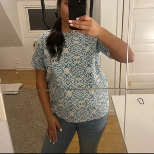 Beige och blå mönstrad t-shirt - T-shirt i beige med blått, detaljerat mönster över hela plagget. Klassisk passform med rund hals och korta ärmar. Materialet är strukturerat och känns lite tjockare än vanlig bomull, vilket ger en unik känsla och look.