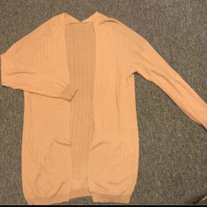 Beige ribbad kofta med lång ärm - Säljer en beige, långärmad kofta med ribbad struktur. Koftan är öppen framtill och har en loose passform som gör den enkel att slänga på över en t-shirt eller topp. Perfekt för dig som gillar neutrala färger och enkel stil.