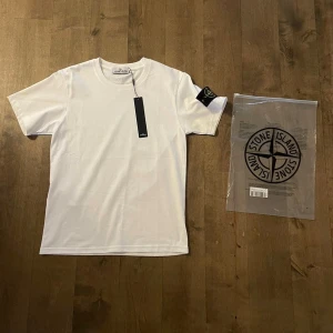 Vit Stone Island t-shirt med patch - Vit t-shirt från Stone Island med klassisk rund hals och korta ärmar. På vänster ärm sitter den ikoniska svarta patchen med broderad gul logga. Materialet är mjuk bomull och passformen är normal. Perfekt för dig som gillar clean och stilren streetwear.