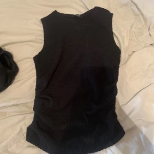 Svart ärmlös linnetopp från Vero Moda - Svart linnetopp från Vero Moda med en cool, strukturerad yta. Modellen är ärmlös och har en rund halsringning. Materialet känns mjukt och har en lätt stretch för skön passform. Perfekt att styla med jeans eller kjol för en enkel och snygg look. Passar även S.