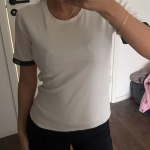 Vit t-shirt med svarta detaljer - Snygg vit t-shirt med rund hals och korta ärmar. Ärmsluten har svarta detaljer med ett diskret hålmönster som ger en cool touch. Passformen är normal och materialet känns mjukt, bomull. Perfekt för dig som gillar stilrena plagg med en twist.