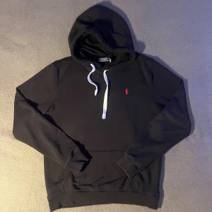 Svart hoodie från Polo Ralph Lauren - Svart hoodie från Polo Ralph Lauren med vit snörning i huvan och klassisk röd broderad logga på bröstet. Tröjan är i storlek large. Tröjan är i ett bra skick och har bara används några gånger. Skriv om du har några funderingar.