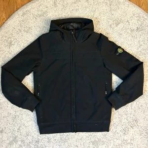 Svart vindjacka från Stone Island - Svart vindjacka från Stone Island med ikonisk patch på ärmen. Jackan har huva, dragkedja framtill och två sidofickor med zip. Materialet är syntet och jackan har en clean, modern look som passar streetwear. Perfekt för dig som gillar stilrena och funktionella plagg.