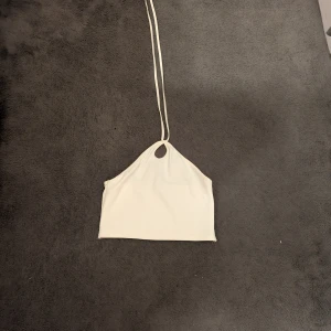 Vit croppad halterneck linnetopp - Säljer en vit croppad linnetopp med halterneck och smala knytband i nacken. Toppen har en enkel och stilren design med en liten öppning vid halsen. 