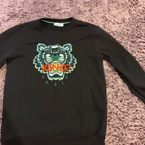 Svart sweatshirt från Kenzo med tiger - Svart sweatshirt från Kenzo med ikonisk broderad tiger på bröstet i grönt, blått och orange. Rund hals, långärmad och normal passform. Materialet är mjuk bomull och tröjan har ribbade muddar vid ärmslut och nederkant. Perfekt för dig som gillar statement-plagg.