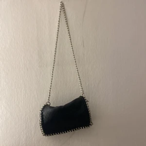 Svart axelväska med kedjedetaljer Tiamo - Cool svart axelväska från Tiamo i skinnimitation med grova silverfärgade kedjor längs kanterna och som axelrem. Väskan har dragkedja både på utsidan och insidan, samt ett stilrent och trendigt utseende som passar perfekt till din outfit.