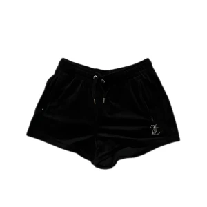 Svarta sammetsshorts Juicy Couture - Svarta sammetsshorts från Juicy Couture med snörning i midjan och fickor. På ena benet finns ett glittrigt JC-emblem och på baksidan står det 'Juicy' i stora strassbokstäver. Perfekta för en trendig och avslappnad look.