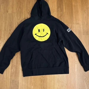Svart hoodie NF - Svart hoodie med stor gul smiley och NF-tryck på magen. Oanvänd 