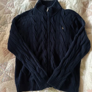 Mörkblå kabelstickad kofta Polo Ralph Lauren - Mörkblå kabelstickad kofta från Polo Ralph Lauren med dragkedja framtill och ribbade muddar. Klassisk broderad logga på bröstet. Hög krage och långärmad modell, perfekt för lager-på-lager under höst och vinter.