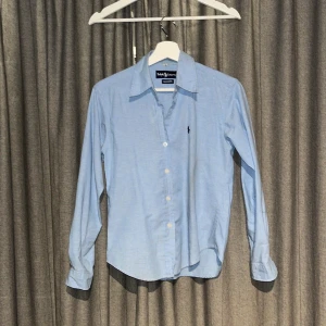 Ljusblå skjorta från Ralph Lauren - Snygg ljusblå skjorta från Ralph Lauren i 100% bomull. Klassisk modell med knappar framtill, lång ärm och broderad logga på bröstet. Perfekt för dig som gillar stilrena och tidlösa plagg med en clean look. En liten defekt är att en knapp har lossnat (se bild 4) men det finns en extra knapp med.