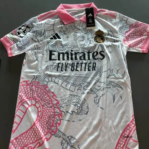 Real Madrid x Adidas vit draktröja - Säljer en Real Madrid fotbollströja med unikt drakmönster i grått och rosa detaljer. Tröjan har korta ärmar, rosa krage och broderat klubbmärke i guld. Materialet är lätt och andas, perfekt för match eller träning. Tröjan är i topp kvalitet.