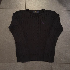 Svart kabelstickad tröja Polo Ralph Lauren - Dam L - Riktigt snygg kabelstickad tröja från Polo Ralph Lauren med lila logga. Tröjan är i gott skick och är i storlek L, osäker om den krympt lite (se bilder för mått). Nypris ligger runt 2200kr