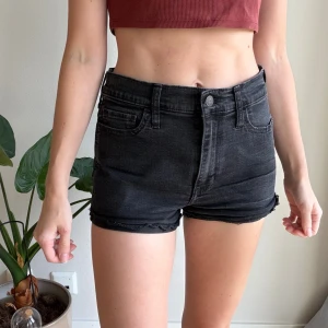 Svarta high rise shorts från Hollister - Snygga svarta jeansshorts från Hollister med hög midja och råa kanter. Klassisk femficksmodell med knapp och dragkedja framtill. Perfekta till sommaren och lätta att matcha med allt. Modellen är kort och sitter tajt över höfterna.