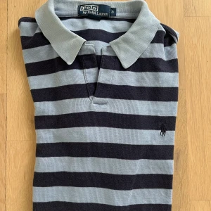 Randig pikétröja från Polo Ralph Lauren - Snygg pikétröja från Polo Ralph Lauren i storlek S. Tröjan har breda horisontella ränder i ljusblått och mörkblått, klassisk krage och korta ärmar. Ikoniska Polo-loggan broderad på bröstet. Perfekt för dig som gillar stilren och sportig look.