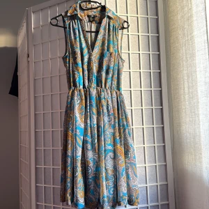 Mönstrad ärmlös klänning från H&M - Säljer en färgstark ärmlös klänning från H&M med paisleyinspirerat mönster i blått, gult och vitt. Klänningen har krage, v-ringning och knappar framtill samt resår i midjan för en snygg siluett. Perfekt för dig som gillar boho-vibe och coola prints.