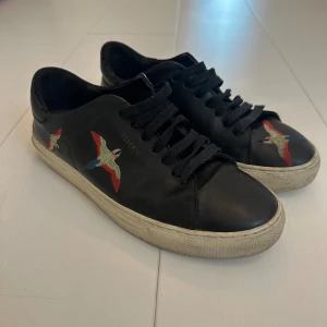 Svarta sneakers från Axel Arigato - Svarta sneakers i skinn från Axel Arigato med broderade fåglar i rött, blått och grönt på sidorna och tån. Klassisk låg modell med svarta snören och vit platt sula. Perfekta för dig som vill ha en stilren sko med unik detalj.
