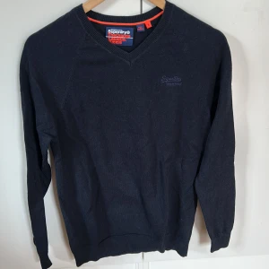 Mörkblå v-ringad tröja Superdry - Snygg mörkblå v-ringad tröja från Superdry Orange Label i storlek S. Tröjan har diskret logga broderad på bröstet och ribbade muddar vid hals, ärmslut och nederkant. Perfekt för dig som gillar stilrena och klassiska plagg.