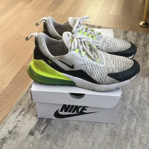 Nike Air Max 270 vita och neongula - Nike Air Max 270 sneakers i vitt mesh med svarta detaljer och neongul häl. Snygg sportig design med synlig Air-enhet i sulan, snörning och rund tå. Perfekta för dig som tänker använda de till gymmet eller till jobbet.