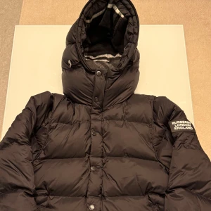 Svart pufferjacka från Burberry - Svart pufferjacka från Burberry med stor huva och tydlig logotyp på ärmen. Jackan har knappar och dragkedja framtill, samt ett rutigt foder i huvan. Perfekt för kalla dagar med sin tjocka och quiltade design.