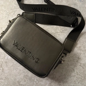 Svart axelväska från Valentino - Snygg svart axelväska från Valentino i skinn med justerbar rem där loggan är tryckt. Väskan har dragkedja upptill, extra fack med dragkedja på baksidan och stilren design. Perfekt för dig som vill ha en trendig och praktisk väska. Ny pris 1200 kr mitt pris 349, ända ”defekterna” är att den har rostat lite vid dragkedjorna. Kontakta mig för bilder på det!