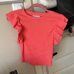 Rosa ribbad topp med volangärmar från Zara - Säljer en superfin rosa topp från Zara i storlek S. Toppen är ribbad och har korta ärmar med snygga volangdetaljer som ger en cool och trendig look. Perfekt att styla med jeans eller kjol för en färgglad vibe.