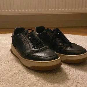 Svarta sneakers från Lyle & Scott - Snygga svarta sneakers från Lyle & Scott i skinn med vit och brun sula. Klassisk design med rund tå, snörning och diskret logga på sidan. Perfekta för dig som gillar stilrena och tidlösa skor med en sportig touch.