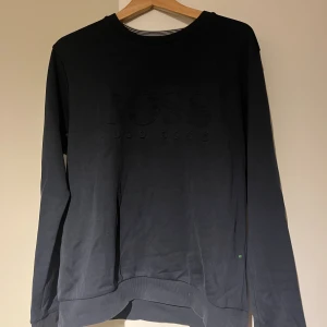 Svart tröja från Hugo Boss - Snygg svart tröja från Hugo Boss med diskret broderad logga på bröstet. Slim fit-modell med rund halsringning och långa ärmar. Tillverkad i mjuk bomull för en bekväm känsla. Perfekt för dig som gillar stilrena och klassiska plagg.