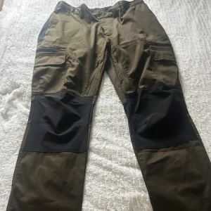 Gröna friluftsbyxor Brokared Dam 50K - Snygga gröna friluftsbyxor från Brokared med svarta förstärkningar över knäna och flera praktiska fickor. Byxorna har dragkedja, knapp och resår i midjan för extra komfort. Perfekta för uteliv och äventyr.