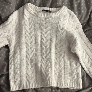 Vit stickad tröja från 157 S - Supermysig vit stickad tröja från 157 med grova flätstickade detaljer och rund hals. Tröjan har lång ärm och ribbade muddar. Perfekt för lager-på-lager under kyliga dagar.