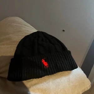 Svart stickad mössa från Polo Ralph Lauren - Svart stickad mössa från Polo Ralph Lauren med klassisk kabelstickning och uppvikt kant. Mössan har en röd broderad logga framtill och är tillverkad i mjukt material som håller dig varm under kalla dagar. Använd endast ett par gånger.