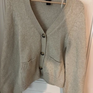 Beige stickad kofta från Lindex - Mysig beige stickad kofta från Lindex i storlek XS. Koftan har djup v-ringning, stora knappar framtill och två fickor. Perfekt att slänga på sig när det är kyligt ute. Enkel och stilren design som passar till det mesta.