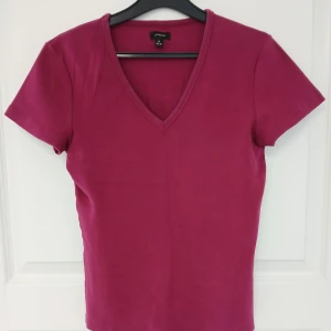 V-ringad topp med korta ärmar - En stilren rosa topp med v-ringning och korta ärmar. Tillverkad i mjuk bomull (95 %) och elastisk elastan (5 %). Perfekt att matcha med jeans eller kjol för en snygg look. Toppen har naturligtvis tvättats. Se bilder för skick och passform. Storlek M enligt lappen men då måttet armhåla till armhåla är cirka 41 cm så stämmer XS-S bättre. Axelsöm till ärmslut är cirka 15 cm. Tvättas i 40 grader.