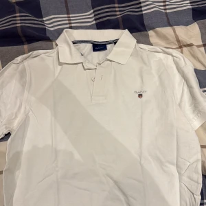 Vit pikétröja från GANT - Vit pikétröja från GANT med klassisk krage och två knappar framtill. Tröjan har korta ärmar och GANT-logga broderad på bröstet. Tillverkad i mjuk bomull som är skön mot huden. Perfekt för dig som gillar stilren och enkel design. (Ej strykt)